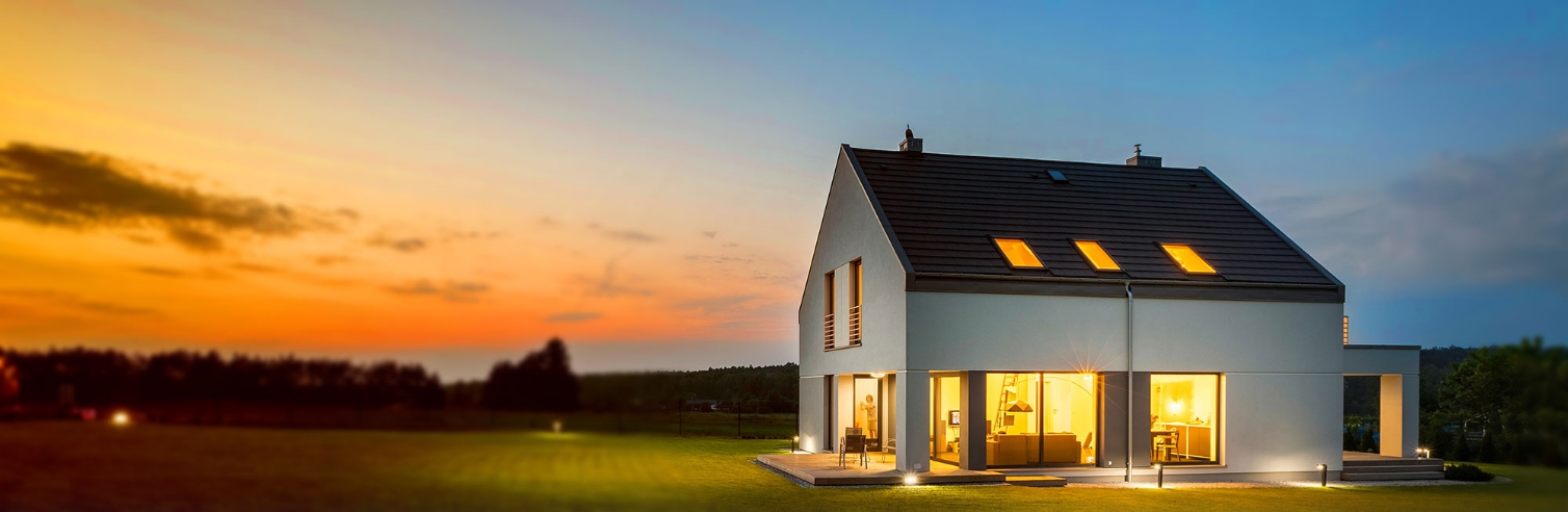 Modernes zweistöckiges Haus mit grauem Ziegeldach und großen Fenstern bei Sonnenuntergang; im Vordergrund eine Rasenfläche, im Hintergrund Bäume und ein farbenfroher Himmel.